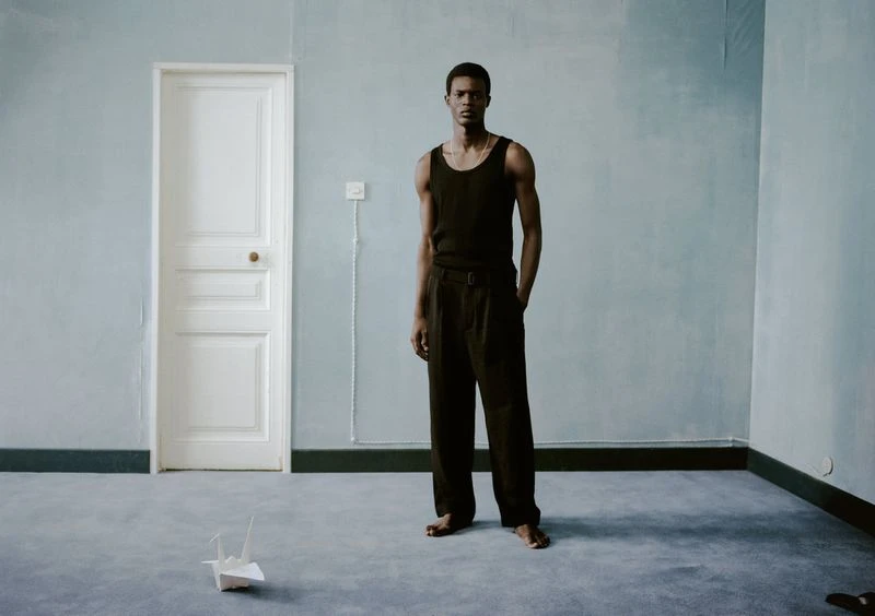 Adult male in black tank top and pants stands in a light blue room with a white door and paper crane on the grey floor.
