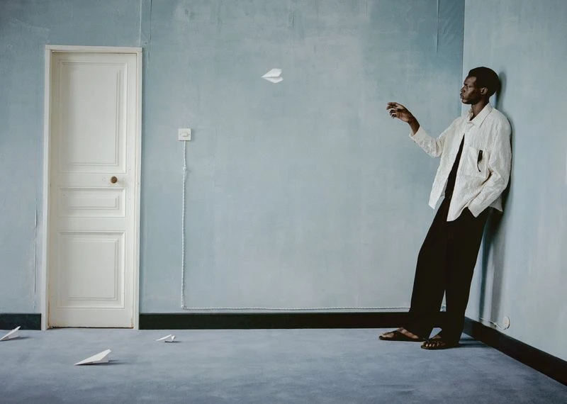Man throwing paper airplane in blue room with light walls, dark trim, and gray carpet; creative lifestyle photography with indoor setting.