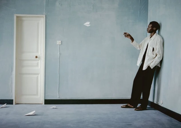 Man throwing paper airplane in blue room with light walls, dark trim, and gray carpet; creative lifestyle photography with indoor setting.