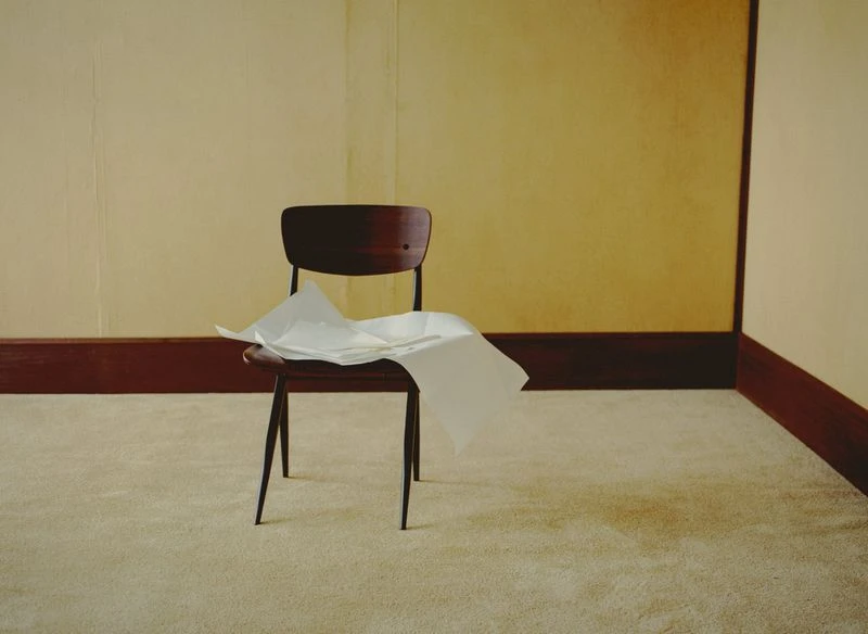 Dark wood chair sits in an empty room with white papers scattered on the seat, against cream and tan walls and beige carpet.