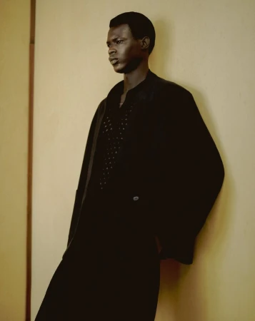 A dark-skinned male model is posing indoors against a light yellow wall. He is wearing a black coat and has a serious facial expression.