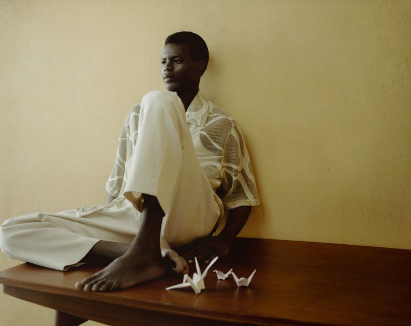 Portrait of a dark-skinned adult man sitting on a dark wooden table in front of a yellow wall with paper origami birds.
