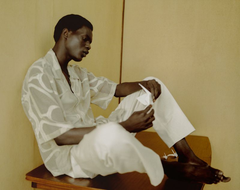 Adult male sits and folds a white paper origami crane. He wears a patterned shirt and white pants. A few origami cranes are visible.