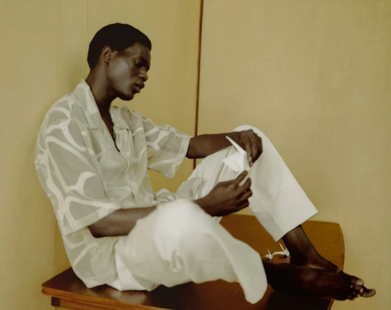 Adult male sits and folds a white paper origami crane. He wears a patterned shirt and white pants. A few origami cranes are visible.