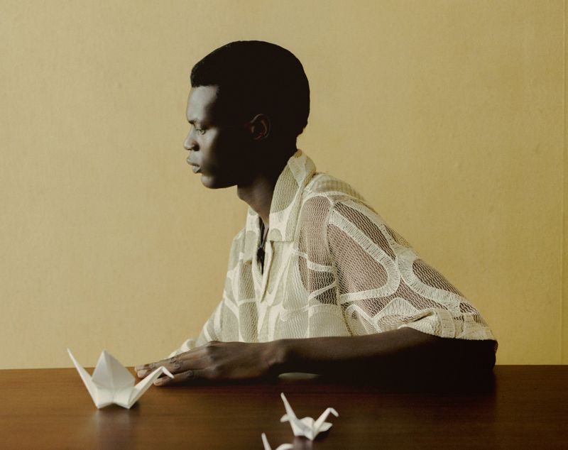 Young man in a patterned shirt sits at a wooden table, creating origami cranes against a muted yellow wall in a natural light setting.