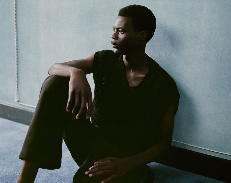 A dark-skinned adult male sits in a dimly lit room, wearing a black knit top and pants, looking off to the side with a serious expression.