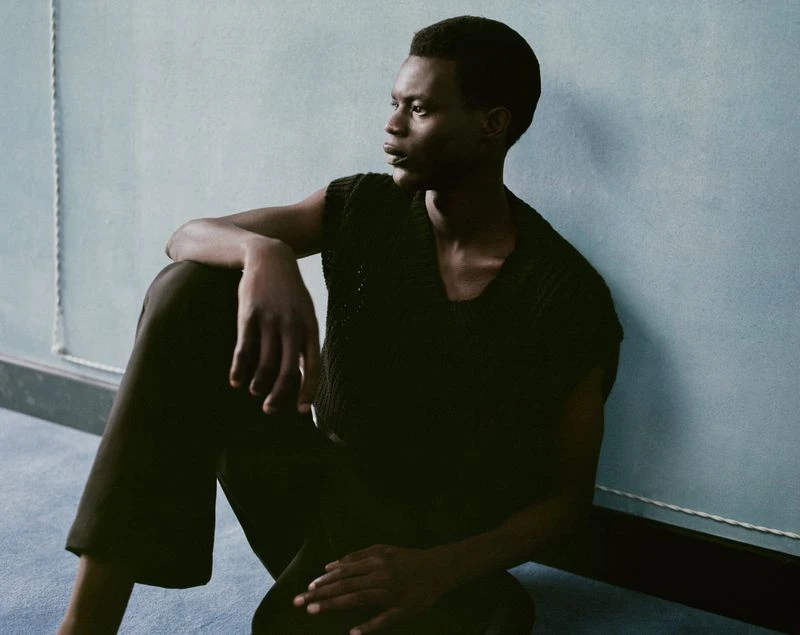 A dark-skinned adult male sits in a dimly lit room, wearing a black knit top and pants, looking off to the side with a serious expression.
