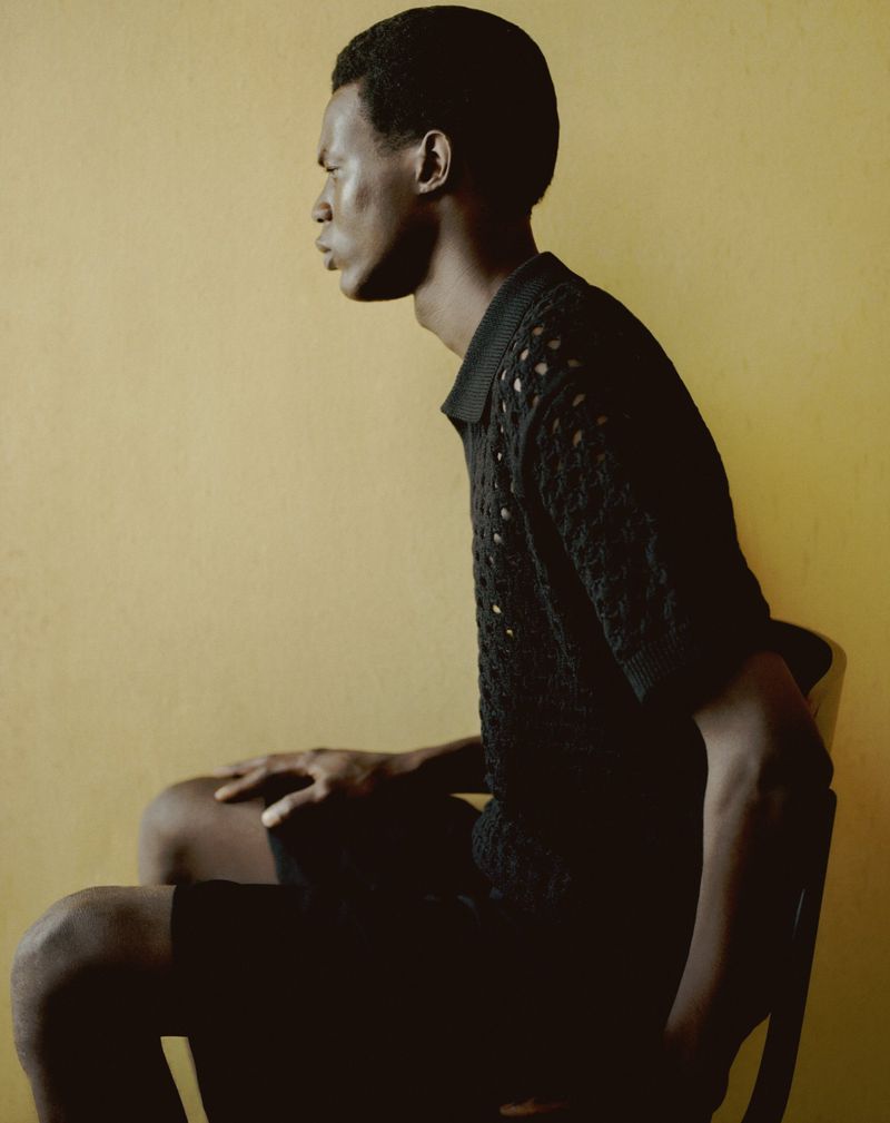 Profile portrait of a Black man seated on a chair against a warm yellow textured wall wearing a black knit polo shirt.