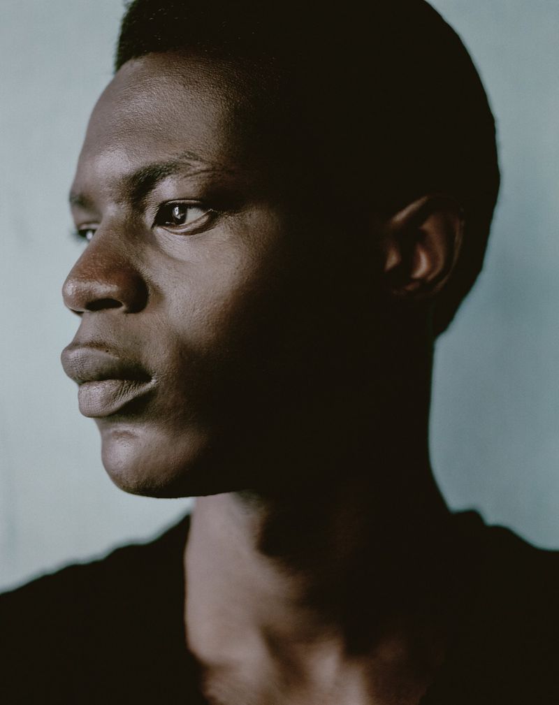 Portrait of an African male in black top with a neutral expression against a light blue background, captured with natural light.