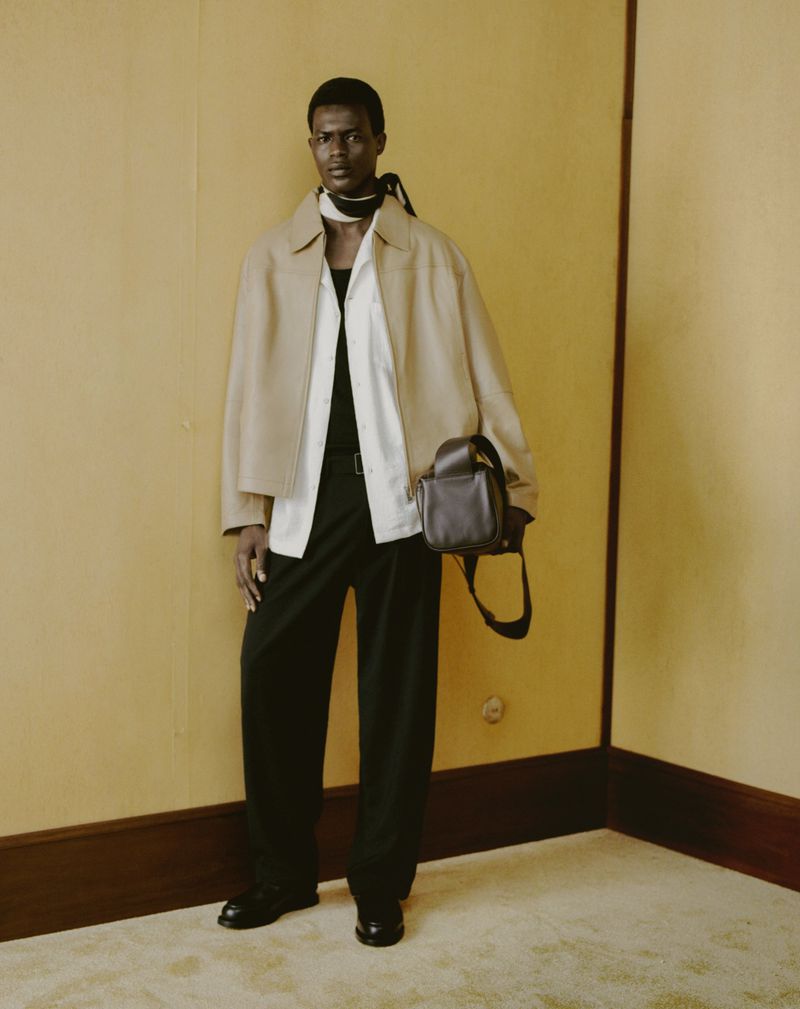 Studio shot of a Black man modeling tan jacket, white shirt, black trousers and shoes, with a dark handbag against a yellow wall.