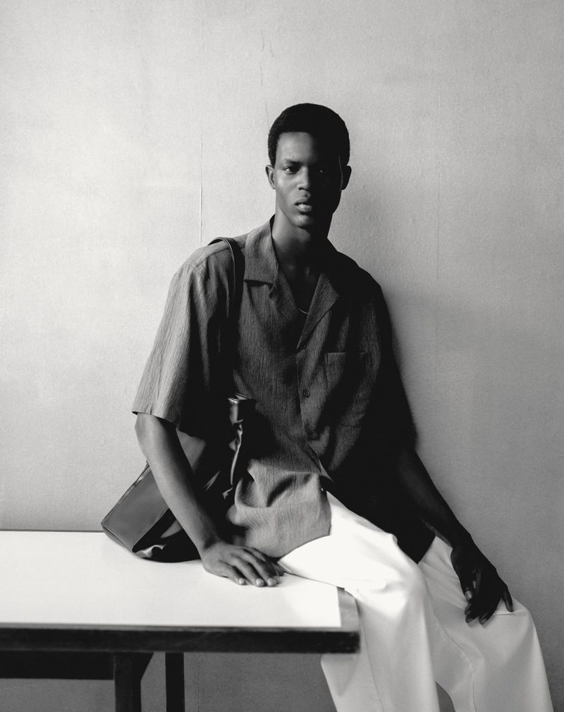 An adult male sits on a white table wearing a button-down shirt and white pants with a bag slung over his shoulder, against a plain background.