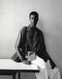 Black man wearing a collared short sleeve shirt and white trousers leaning against a wall.