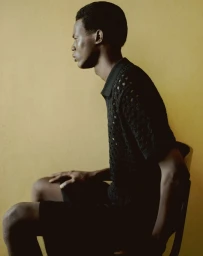 Profile portrait of a Black man seated on a chair against a warm yellow textured wall wearing a black knit polo shirt.