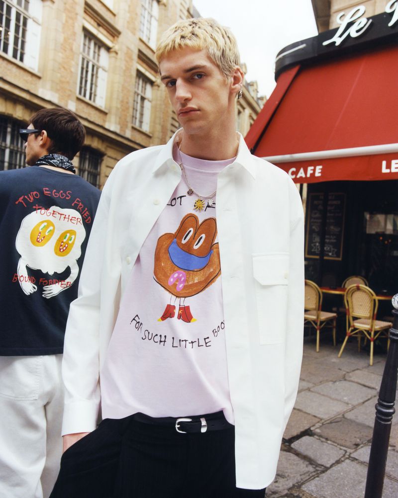 Two men in an urban setting. One man is in focus wearing a white button up shirt over a pink shirt. Cafe is in the background.