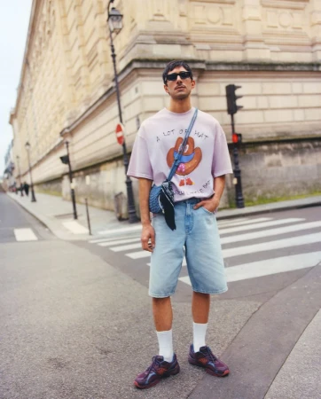 An adult male stands in an urban setting wearing a graphic tee, light wash shorts, socks and sneakers, and a cross-body bag over his shoulder.