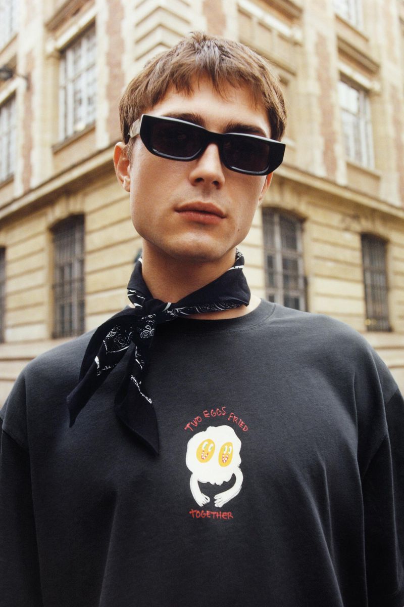 A young man with a black bandana and sunglasses poses in front of a beige building. He wears a black sweatshirt with a white egg graphic.