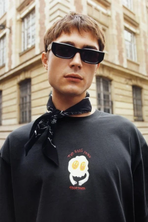 A young man with a black bandana and sunglasses poses in front of a beige building. He wears a black sweatshirt with a white egg graphic.