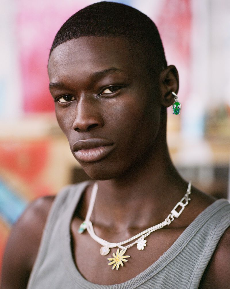 Close-up of a dark-skinned adult male wearing jewelry: a silver chain necklace with charms and a frog earring, against a colorful background.