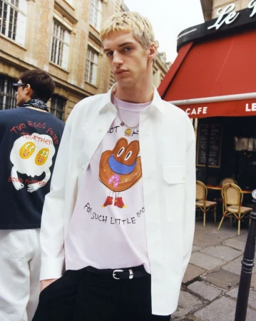 Blonde male model wearing a white button-down shirt and graphic t-shirt standing outside a cafe.