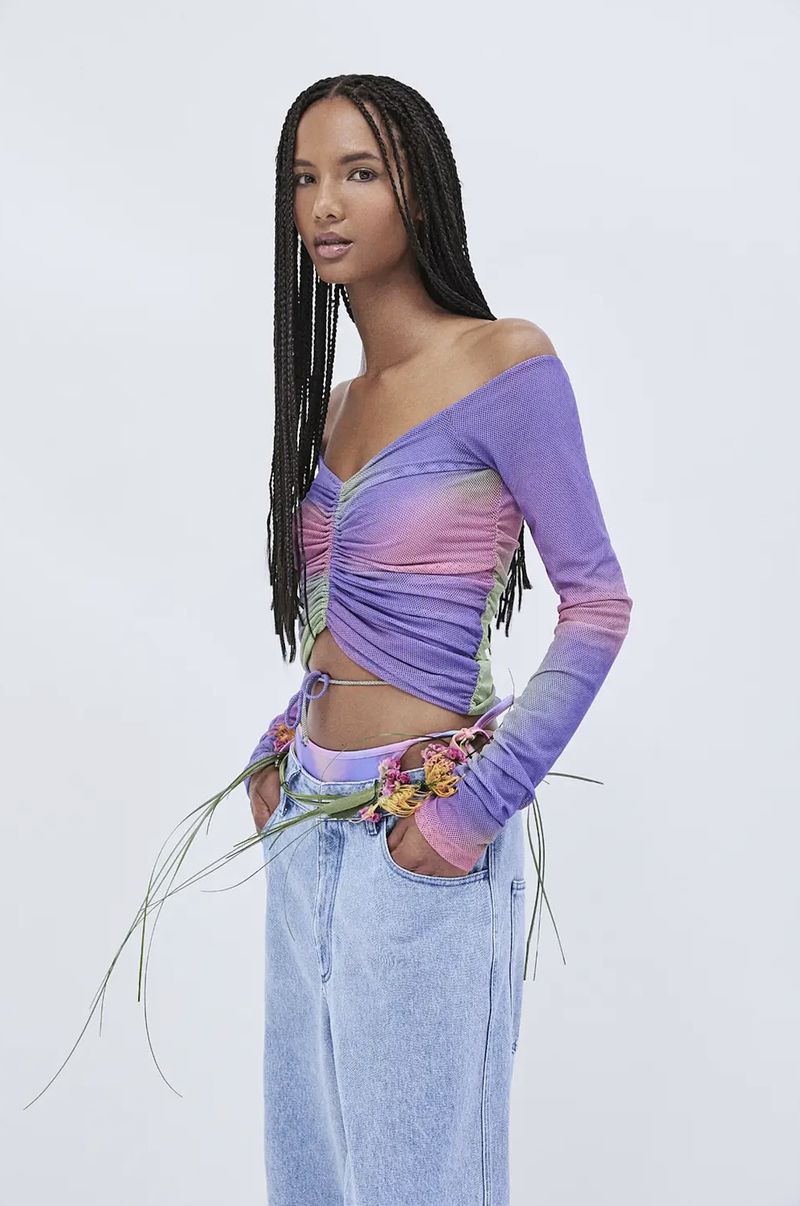 Woman poses in a studio wearing a purple and pink off-the-shoulder top and light blue jeans, accessorized with flowers. Fashion shot.