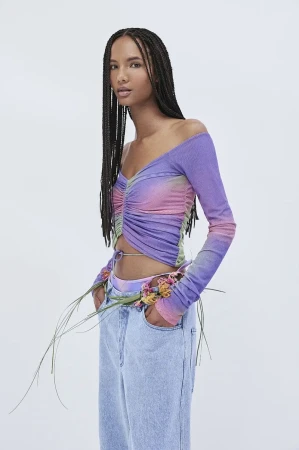 Woman poses in a studio wearing a purple and pink off-the-shoulder top and light blue jeans, accessorized with flowers. Fashion shot.