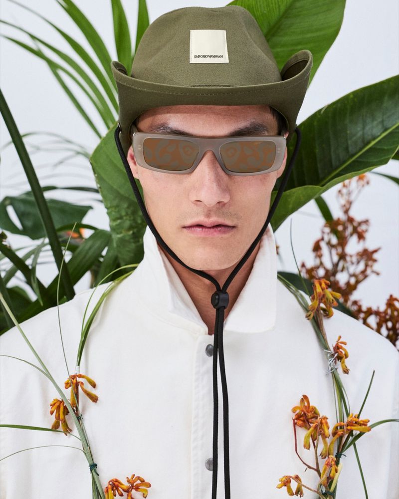 Portrait of a stylish adult male wearing a white jacket, olive hat, and sunglasses, posing in front of tropical plants.