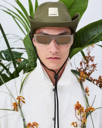 Portrait of a stylish adult male wearing a white jacket, olive hat, and sunglasses, posing in front of tropical plants.