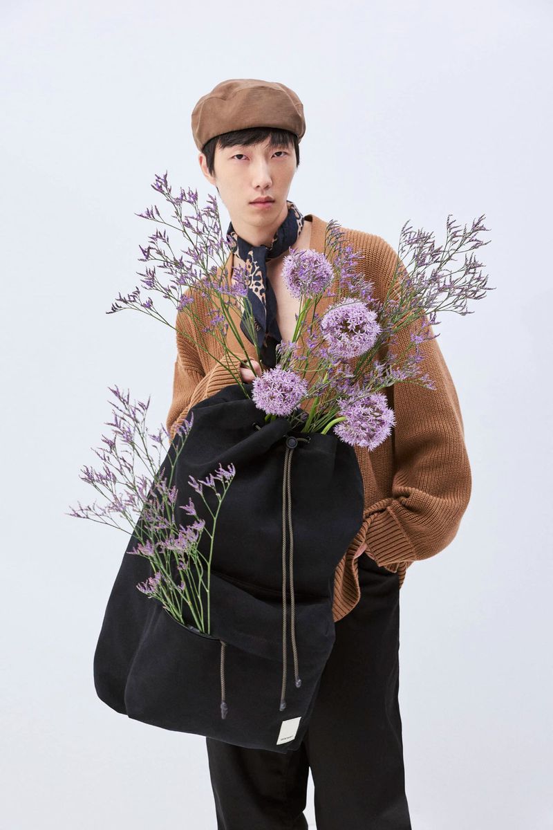 Portrait of a stylish Asian man in beret and cardigan holding a black drawstring bag filled with purple allium flowers on white.