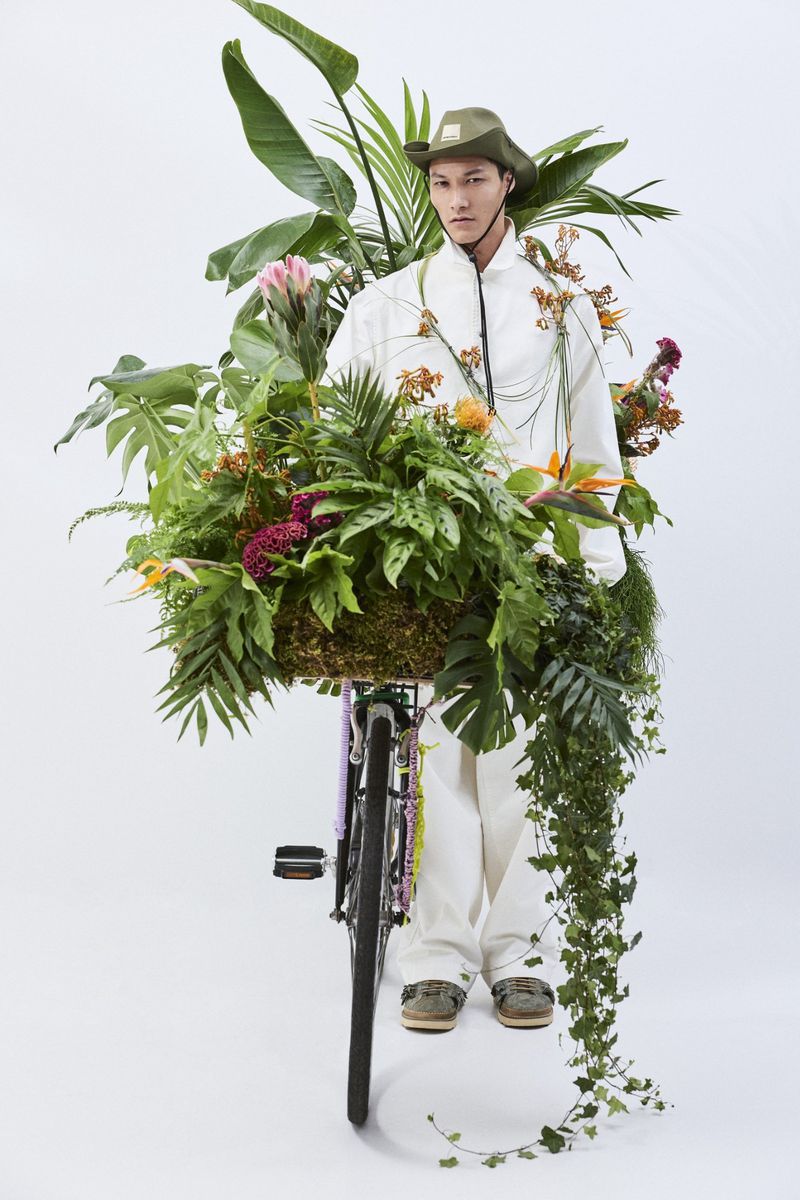 A person in a white outfit and olive hat stands behind a bicycle covered in lush tropical plants against a plain white background.