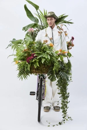 A person in a white outfit and olive hat stands behind a bicycle covered in lush tropical plants against a plain white background.