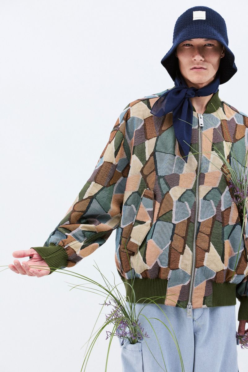 Person in patterned jacket and blue bucket hat posing in a white studio setting. Fashion shot with plants and light blue pants.