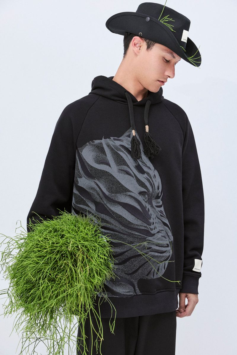 Adult male in a studio setting wearing a black hoodie with a tiger graphic and a cowboy hat holds a green plant.