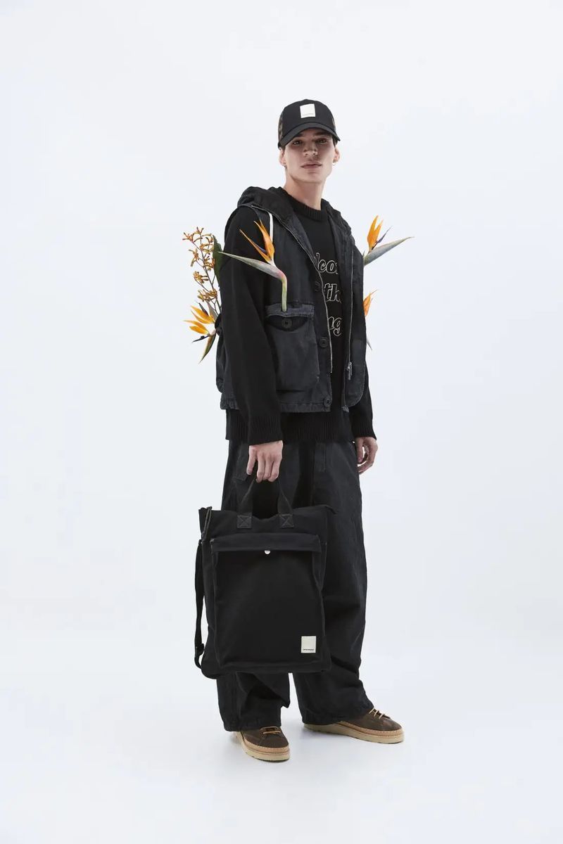 Man in streetwear fashion wearing a vest with bird of paradise flowers, holding a black tote bag in a white photo studio setting.