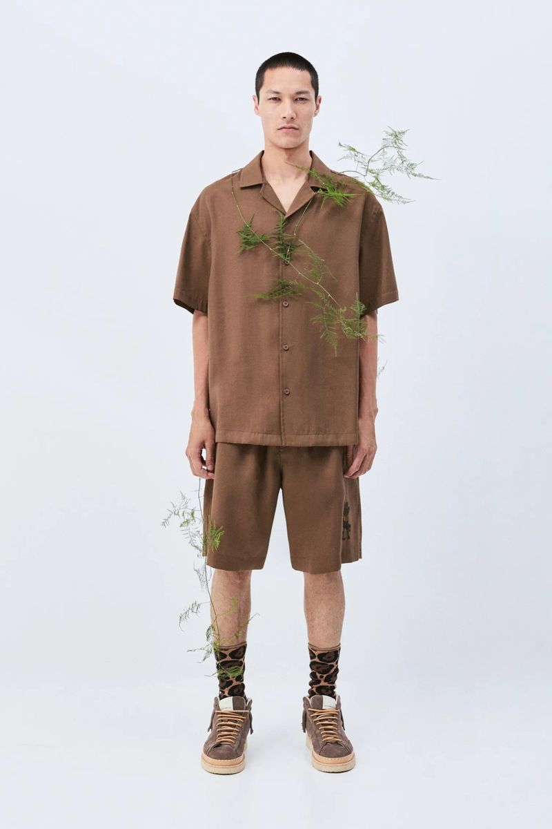 A slender man poses in a brown collared short-sleeve shirt and matching shorts, complemented by patterned socks and brown sneakers in a studio.