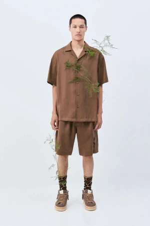 A slender man poses in a brown collared short-sleeve shirt and matching shorts, complemented by patterned socks and brown sneakers in a studio.