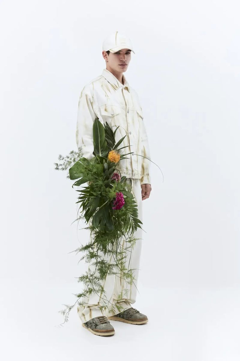 Male fashion model poses in a cream-colored outfit with a floral arrangement against a stark white backdrop in a studio setting.