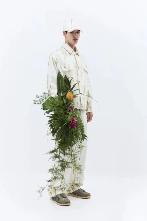 Male fashion model poses in a cream-colored outfit with a floral arrangement against a stark white backdrop in a studio setting.
