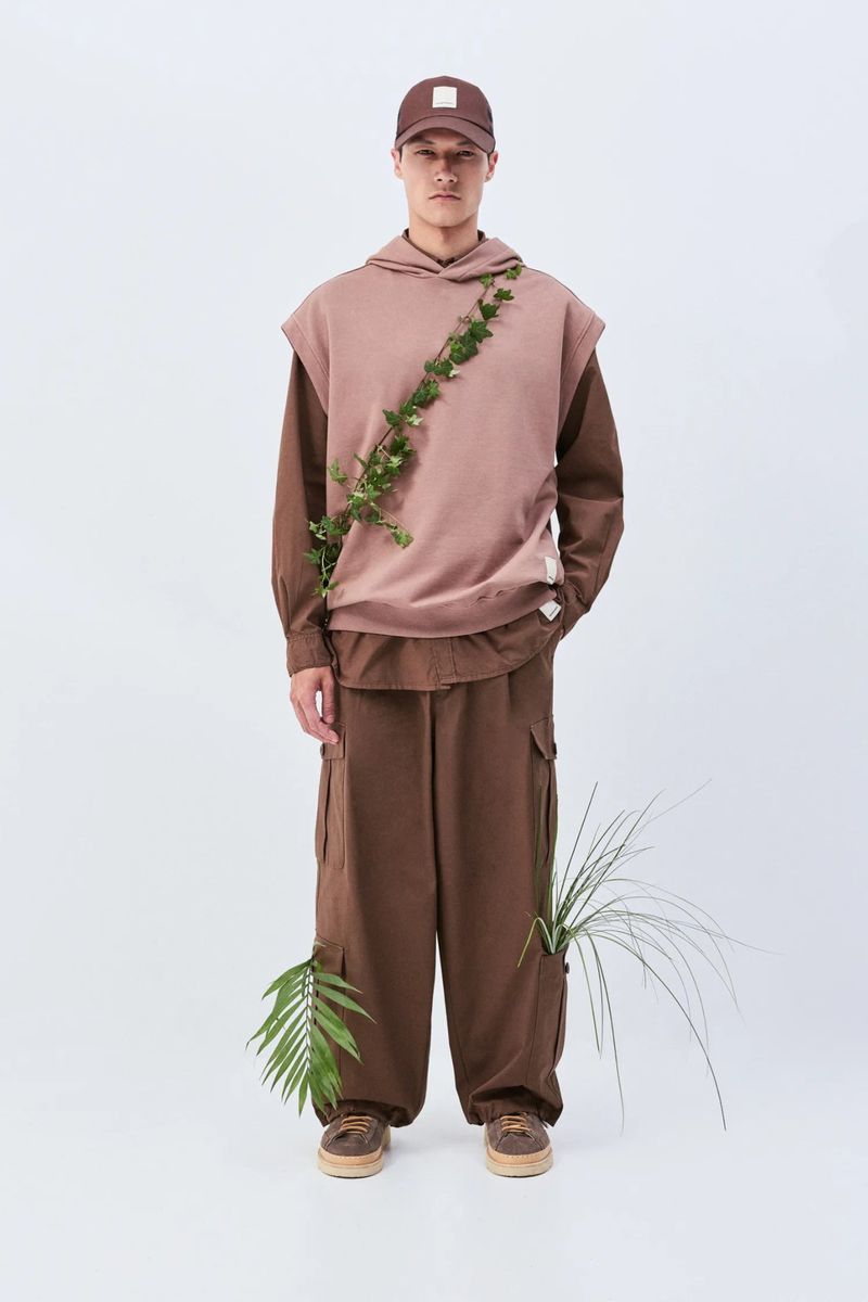 Full-length shot of a male model in a brown hooded vest, brown cargo pants, baseball cap, accessorized with green leafy vines in a white studio.