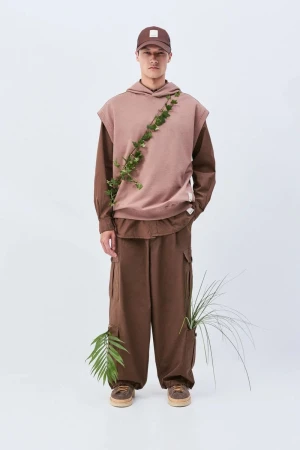 Full-length shot of a male model in a brown hooded vest, brown cargo pants, baseball cap, accessorized with green leafy vines in a white studio.