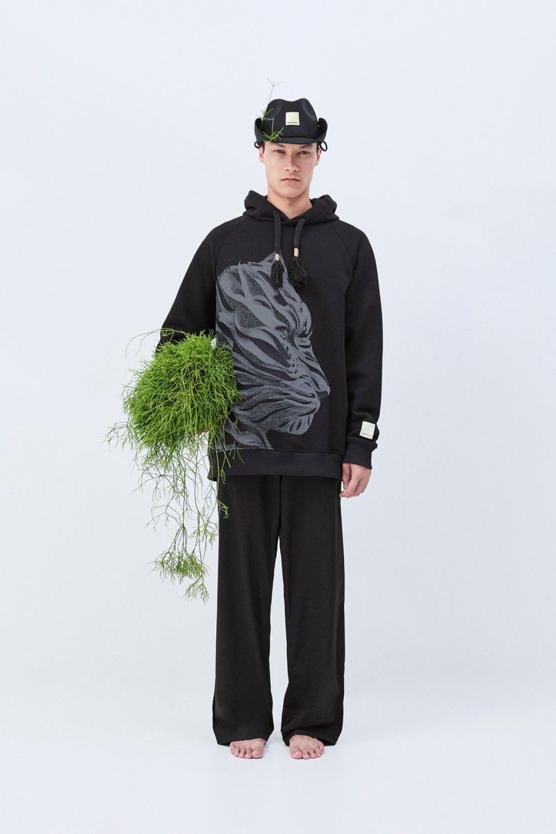 An adult man stands in a studio, wearing a black hooded sweatshirt with a tiger graphic, matching pants, a hat, and holding greenery.