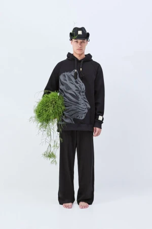 An adult man stands in a studio, wearing a black hooded sweatshirt with a tiger graphic, matching pants, a hat, and holding greenery.