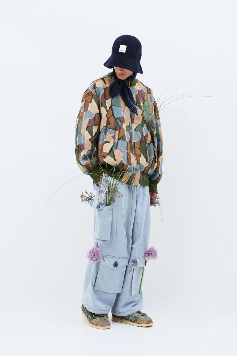 Person in patterned jacket, light blue pants, and hat with flowers, standing against a white backdrop in a fashion-editorial style.