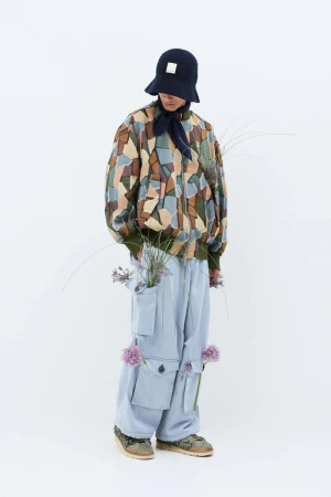 Person in patterned jacket, light blue pants, and hat with flowers, standing against a white backdrop in a fashion-editorial style.