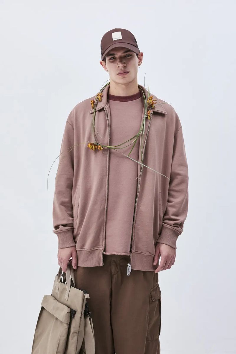 A young adult male stands in a photo studio wearing casual clothing, a baseball cap, and a jacket, holding a tote bag.