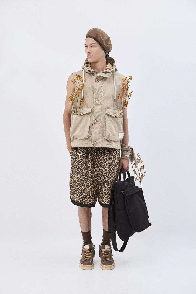 Full-length studio shot of a male model in a sleeveless coat, patterned shorts, and beret, holding a black backpack against white.