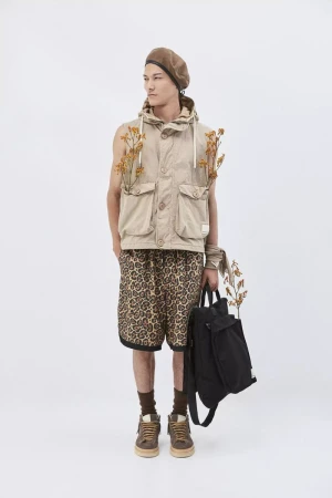 Full-length studio shot of a male model in a sleeveless coat, patterned shorts, and beret, holding a black backpack against white.
