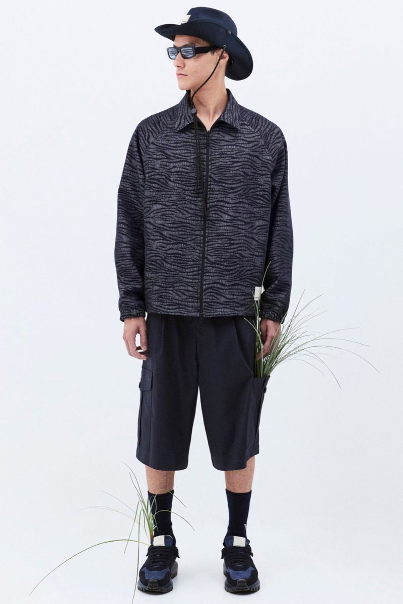 Stylish adult male in a patterned jacket, navy shorts, hat, and sunglasses, standing indoors in a fashion pose against a white background.