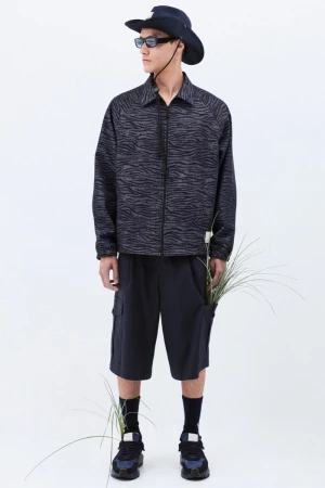 Stylish adult male in a patterned jacket, navy shorts, hat, and sunglasses, standing indoors in a fashion pose against a white background.