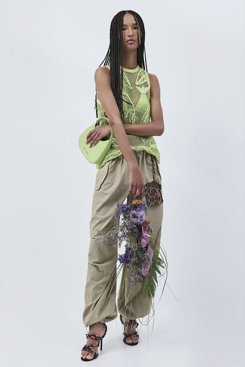 Full shot of an adult female model in a neon green mesh top, beige pants, and strappy heels, holding a green purse and bouquet against white.