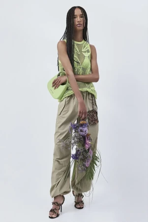Full shot of an adult female model in a neon green mesh top, beige pants, and strappy heels, holding a green purse and bouquet against white.